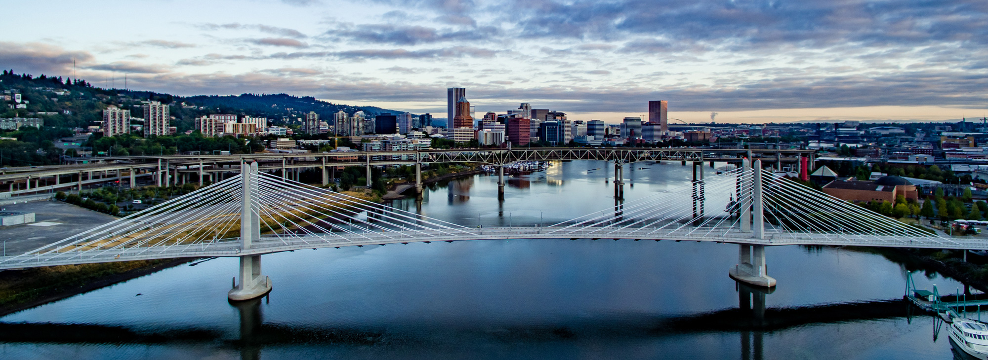 A cable-stayed bridge spans a calm river, with a city skyline featuring tall buildings and a cloudy sky in the background, creating an urban landscape.