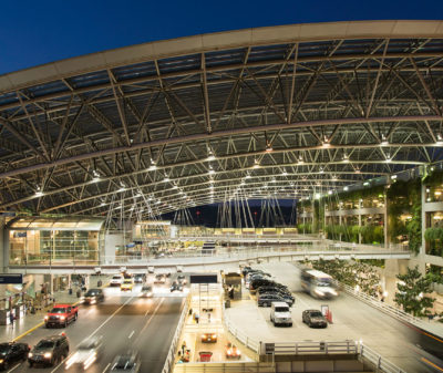 A large, illuminated airport terminal with an arched glass and steel roof, bustling with moving vehicles below. Adjacent, a multi-level parking garage displays parked cars and vertical greenery.