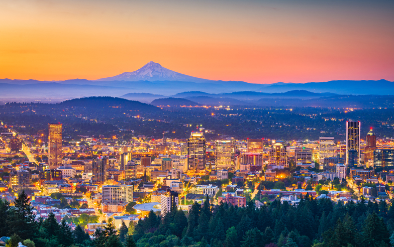 City skyline illuminated at dusk, buildings glowing with lights against a backdrop of distant mountains. The sky transitions from orange to blue, creating a serene and vibrant urban landscape.