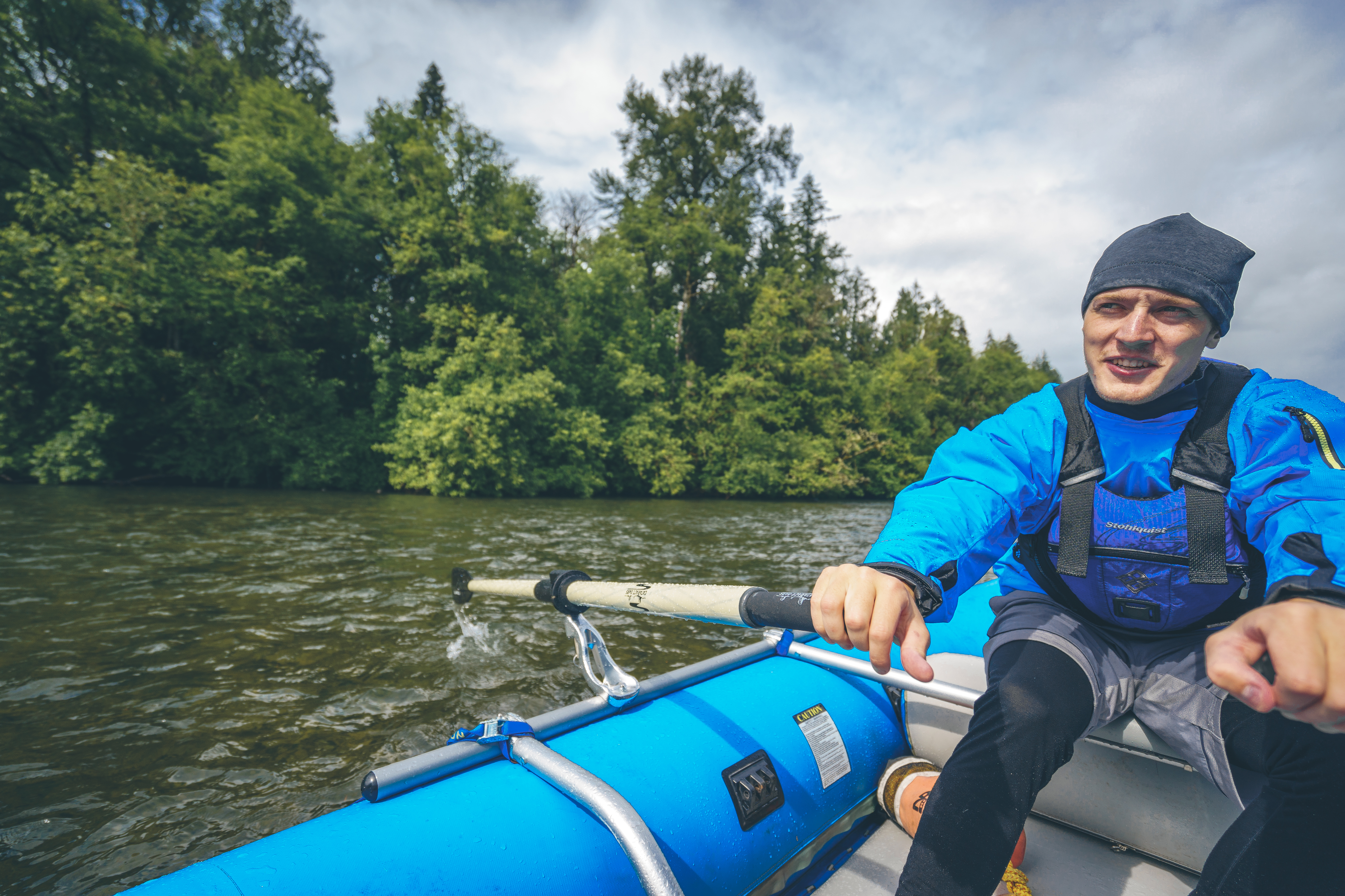 A person in a blue jacket and hat is vigorously rowing an inflatable boat on a river, surrounded by lush, green trees under a partly cloudy sky.