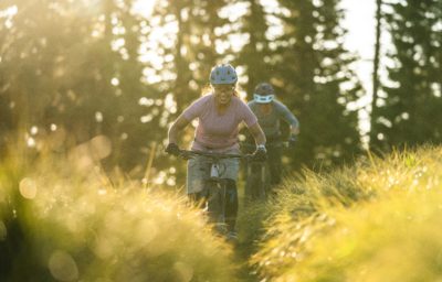 Two women mountain biking on a grassy, sunny biking trail.