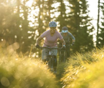 Two women mountain biking on a grassy, sunny biking trail.