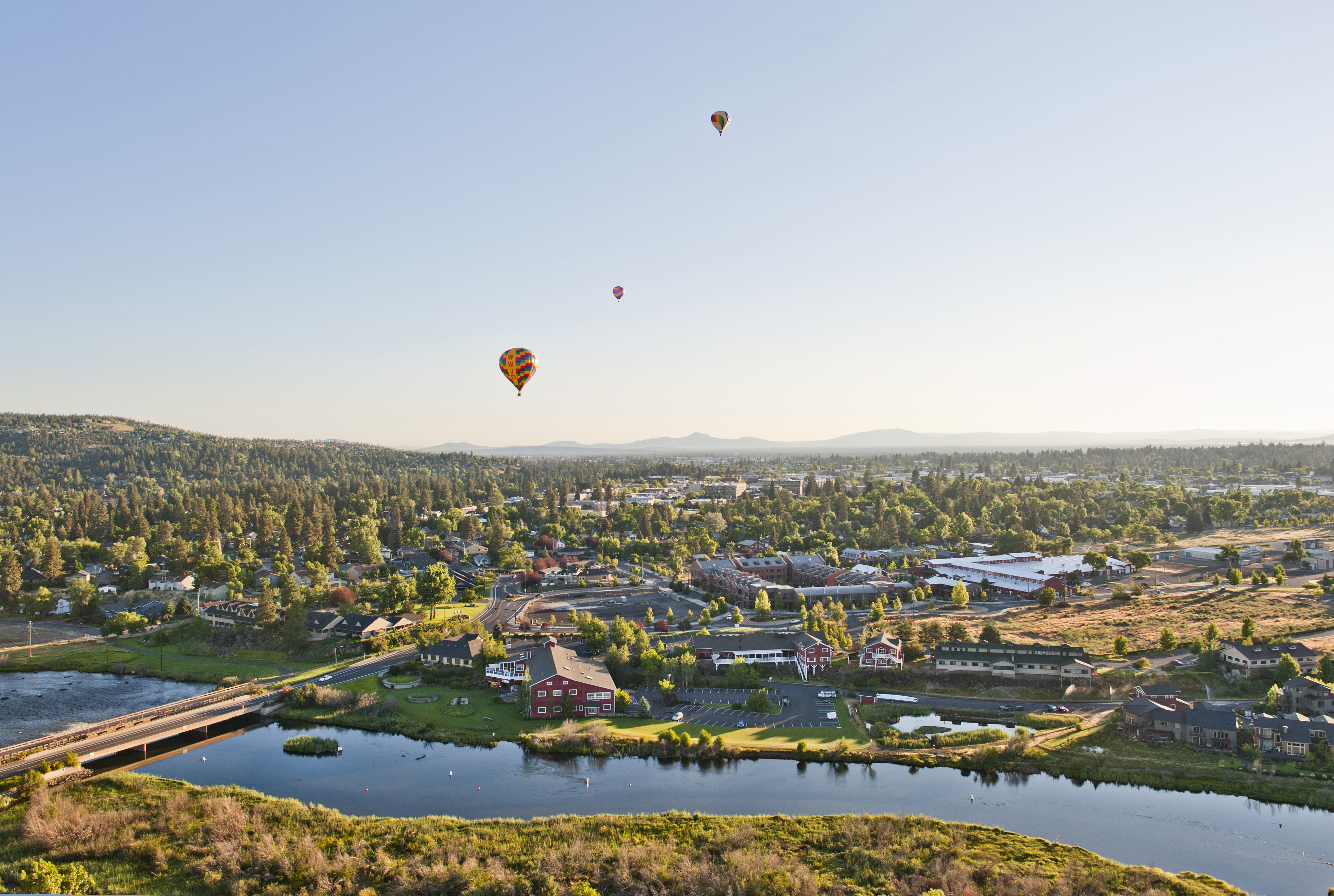 Aerial view of Bend, the Deschutes River and Old Mill district with greenery and a blue sky with three hot air balloons floating down.