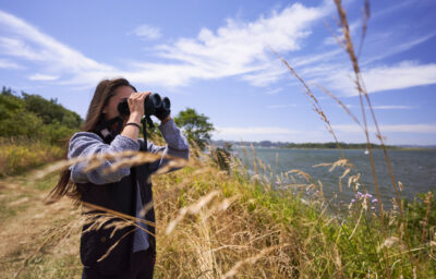 Woman looking out at the coast with binoculars, surrounded by beach grass.