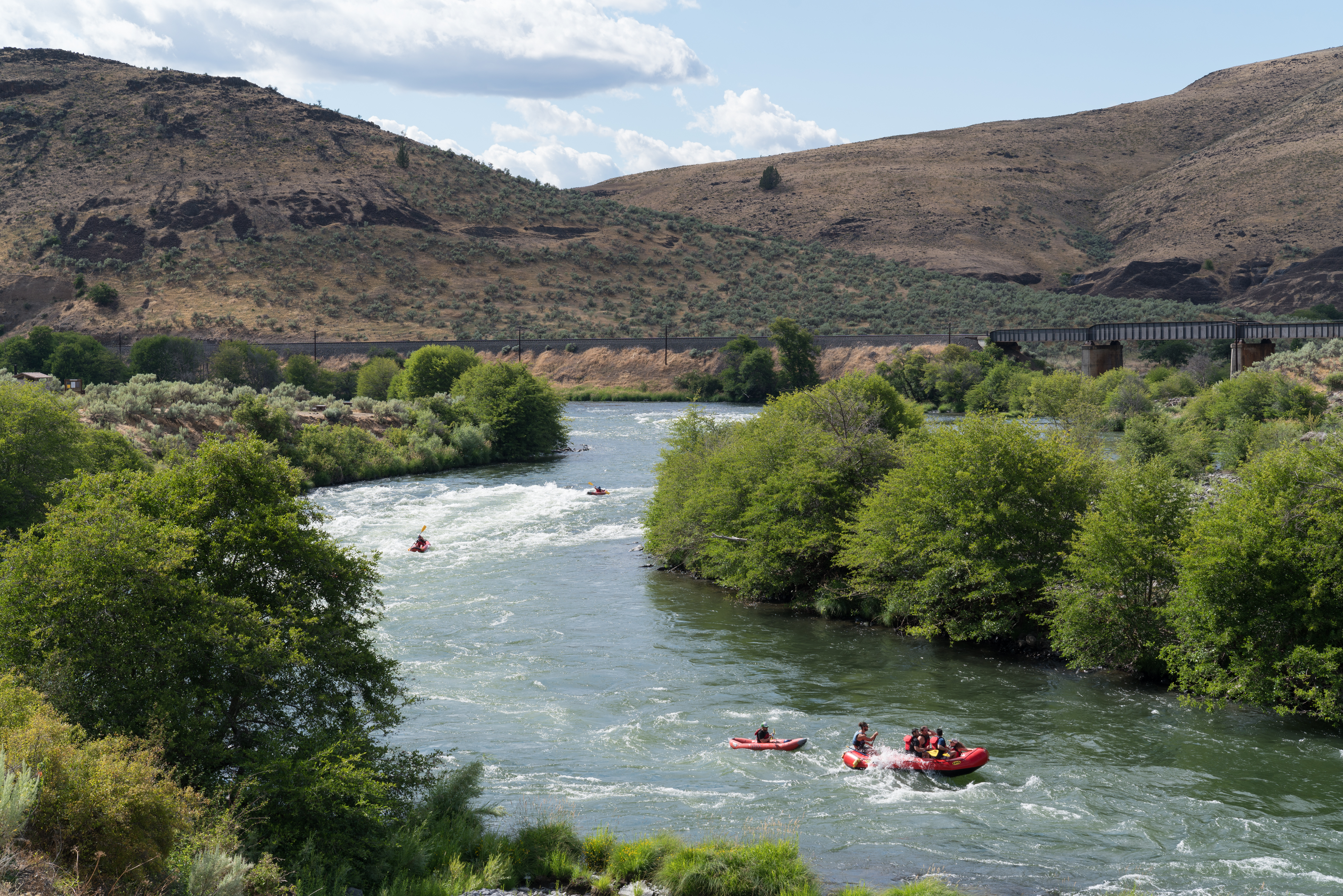 Kayaks are navigating a winding, white-water river surrounded by lush greenery and hills. A railroad bridge spans across the scene in the background, connecting the hilly terrain.