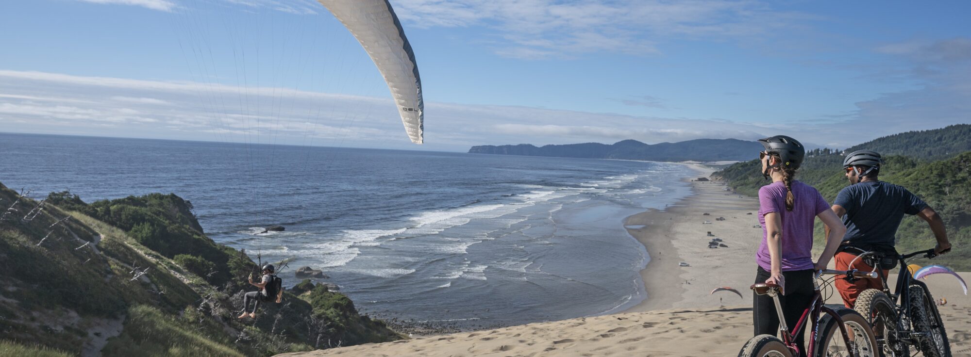 Two cyclists riding through the sand with a view of a hang glider and the ocean in Pacific City.