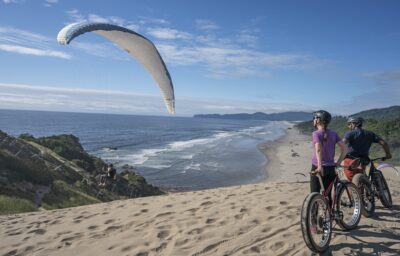 Two cyclists riding through the sand with a view of a hang glider and the ocean in Pacific City.