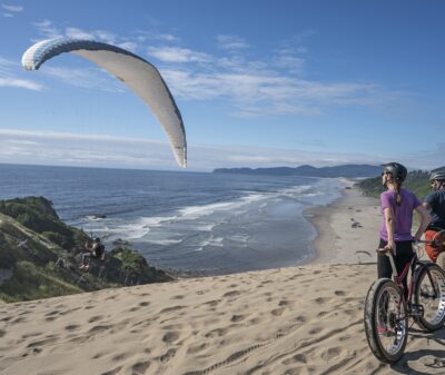 Two cyclists riding through the sand with a view of a hang glider and the ocean in Pacific City.