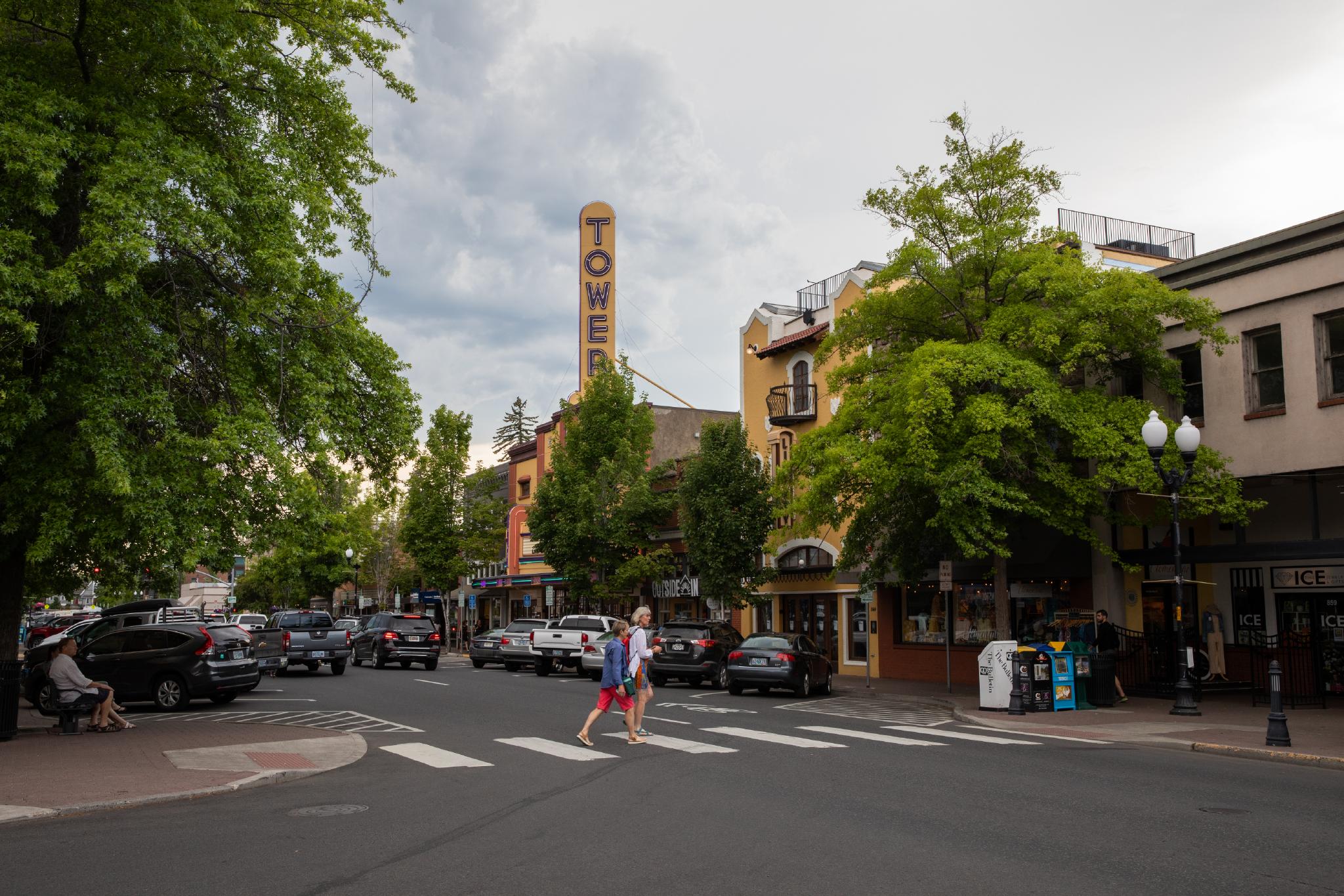 A bustling street features cars and pedestrians under lush green trees. A vertical sign reads "TOWER" on a historic theater building, surrounded by colorful storefronts and overcast skies.