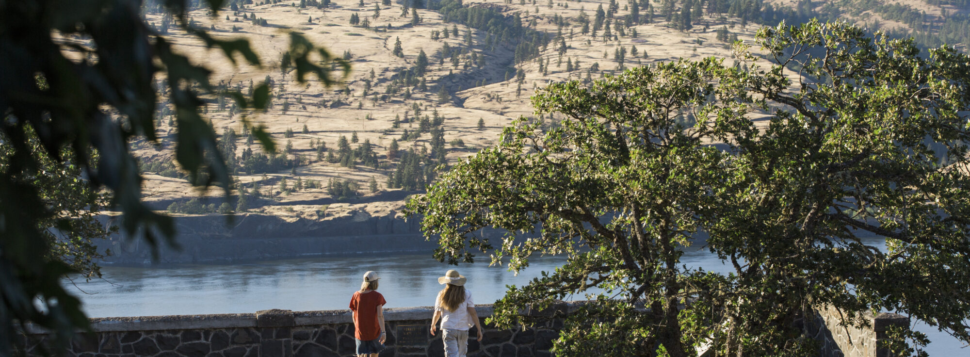Two people walk along a path toward a stone wall, overlooking a river. The scene is framed by trees, with rolling hills in the background under a clear sky.