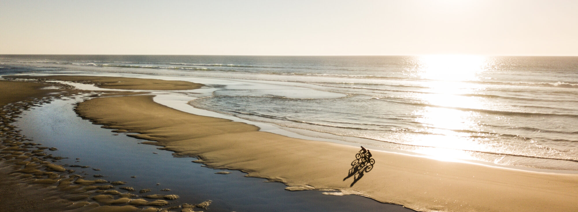 A person rides a bike along a sandy beach at low tide, with the sun shining brightly over the sea, creating reflections on the wet sand.