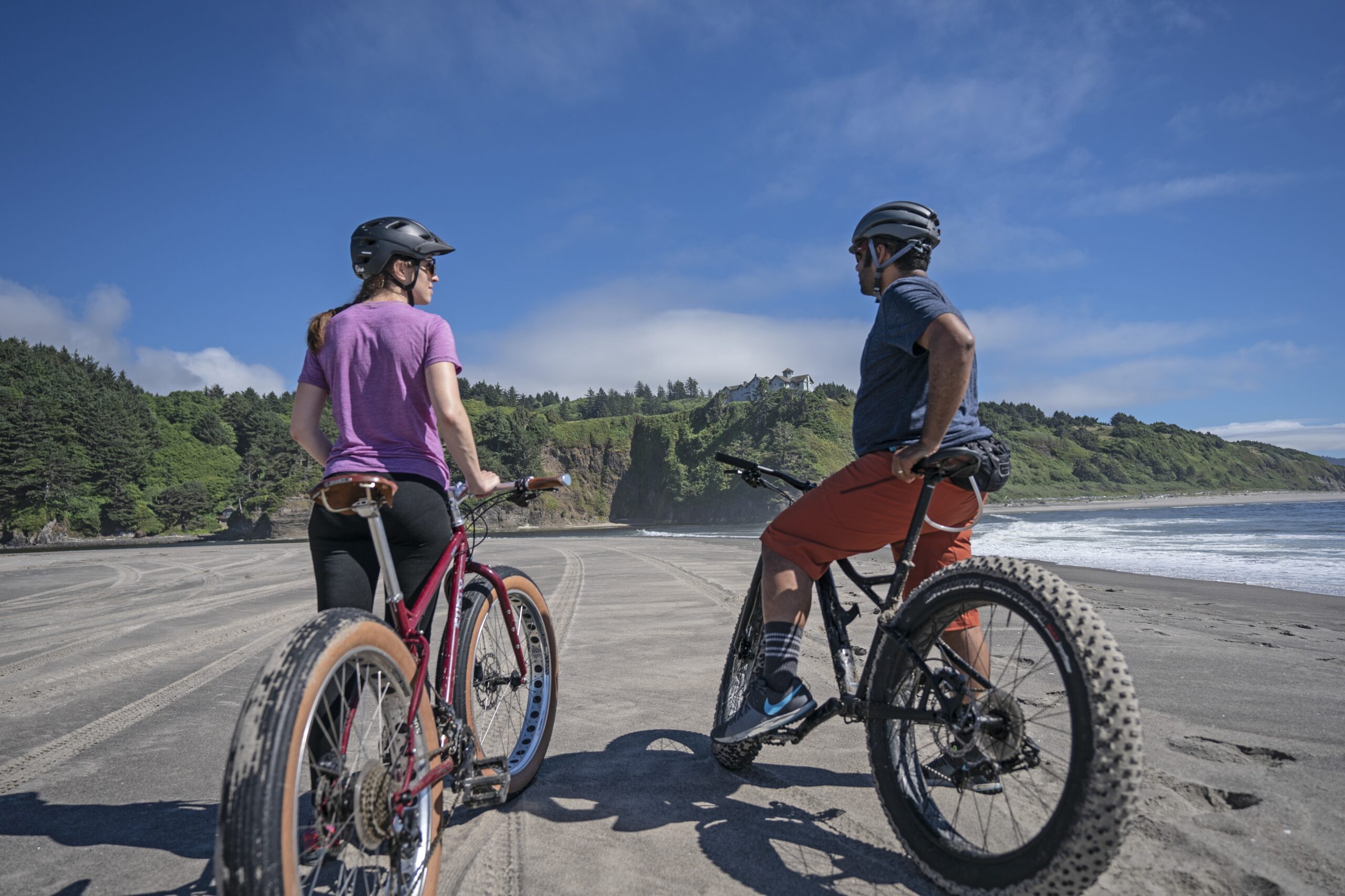Two people on fat bikes pause on a sandy beach, looking toward the ocean. They are surrounded by lush greenery and a clear blue sky.
