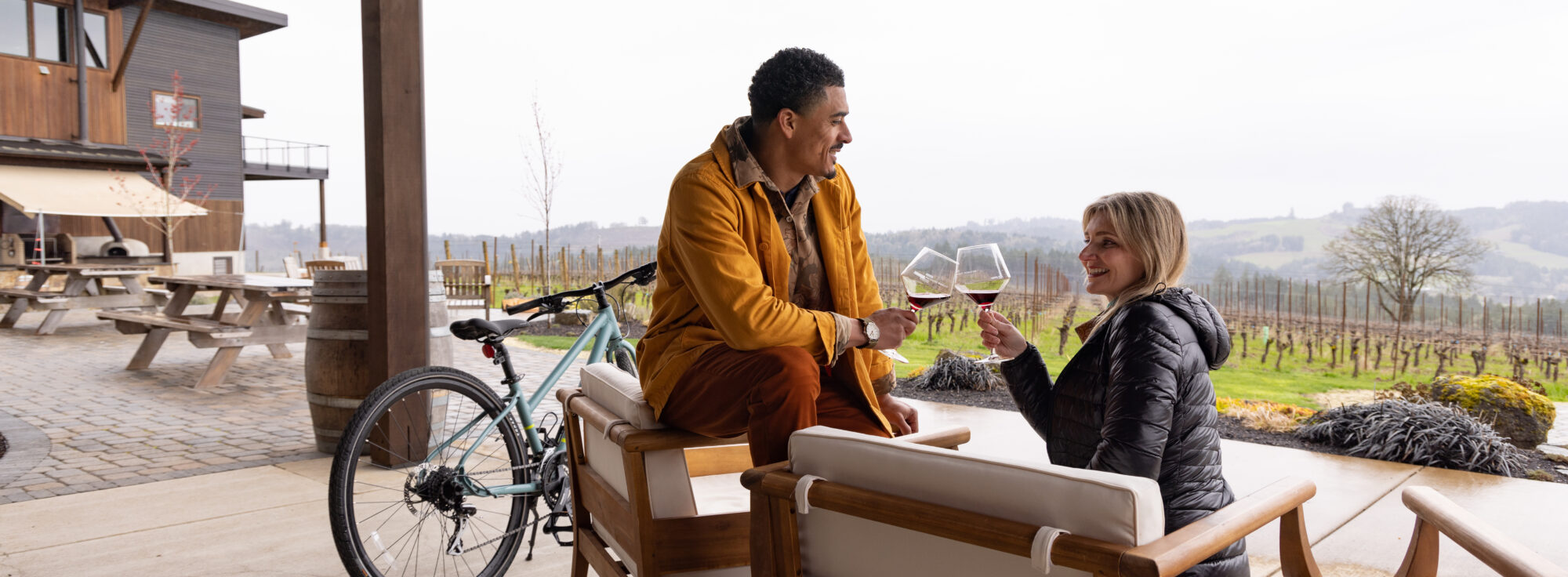 A man and a woman clink wine glasses on a patio overlooking a vineyard. A bicycle leans against a chair, and a modern building sits adjacent to picnic tables.