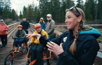 A guide leading a group of six people across a wooden dock. Group of people are on bikes, accessible bikes and standing.