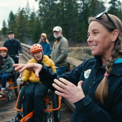 A guide leading a group of six people across a wooden dock. Group of people are on bikes, accessible bikes and standing.