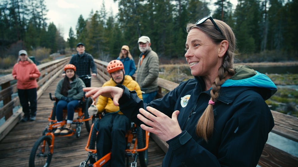 A guide leading a group of six people across a wooden dock. Group of people are on bikes, accessible bikes and standing.