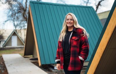 A person in a red plaid jacket stands smiling, hands in pockets, near A-frame wooden structures with green roofs. The setting appears outdoors with bare trees in the background.