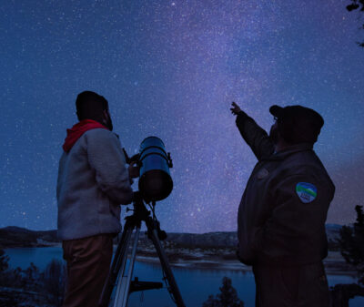 Two people stand by a telescope; one points skywards under a star-filled night. They are near a lake with trees silhouetted, suggesting a remote, natural setting.