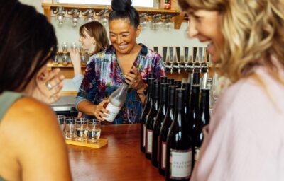 A person smiles while holding a wine bottle behind a bar counter. Several wine bottles and tasting glasses are displayed in front. Other people chat and enjoy the bright, casual setting.