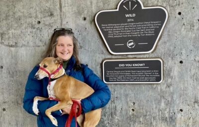 A woman in a blue jacket holds a small brown dog, standing beside a wall with informational plaques. The plaques read:    "WILD 2014   Reese Witherspoon played Oregon author Cheryl Strayed in the memoir adaptation and Pacific Crest Trail filming scenes here also doubled for the Sierra Nevada. Wild contained more Oregon film sites than any prior Oscar-nominated film. Oregon film history: Gus Van Sant directed Uma Thurman in Even Cowgirls Get The Blues (1994), adapted from the novel by Tom Robbins."    "DID YOU KNOW?   Central Oregon and Smith Rock have long been a draw for Hollywood filmmakers. In fact, Oregon’s first two movies were made in the High Desert. Filmed in the mountaintains, in and near Klamath proto-western The Indian Tomb (1914), and in the high desert, Florence Turner’s movie, Love’s Folly (fall 1914). More recently: Rooster Cogburn (1975), Fire in the Sky (1993), and Swordfish (2001)."