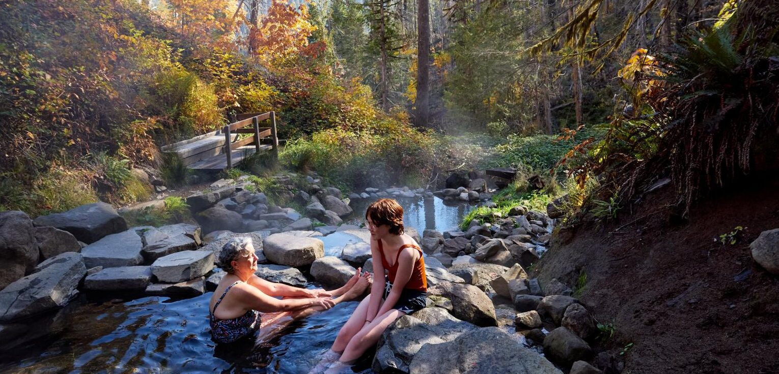 Two people sitting in a hotspring surrounded by rocks, trees and sunshine.