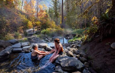 Two people sitting in a hotspring surrounded by rocks, trees and sunshine.