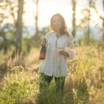Woman in a white blouse with long brown hair standing in a sun filled field holding a bottle of wine.