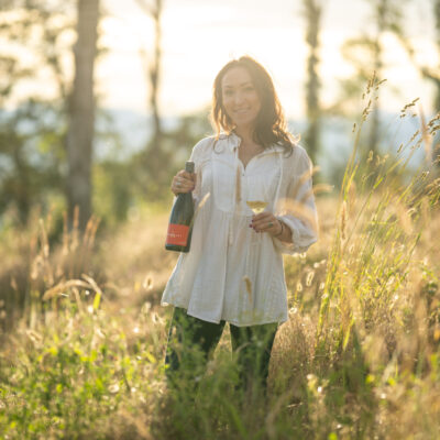 Woman in a white blouse with long brown hair standing in a sun filled field holding a bottle of wine.
