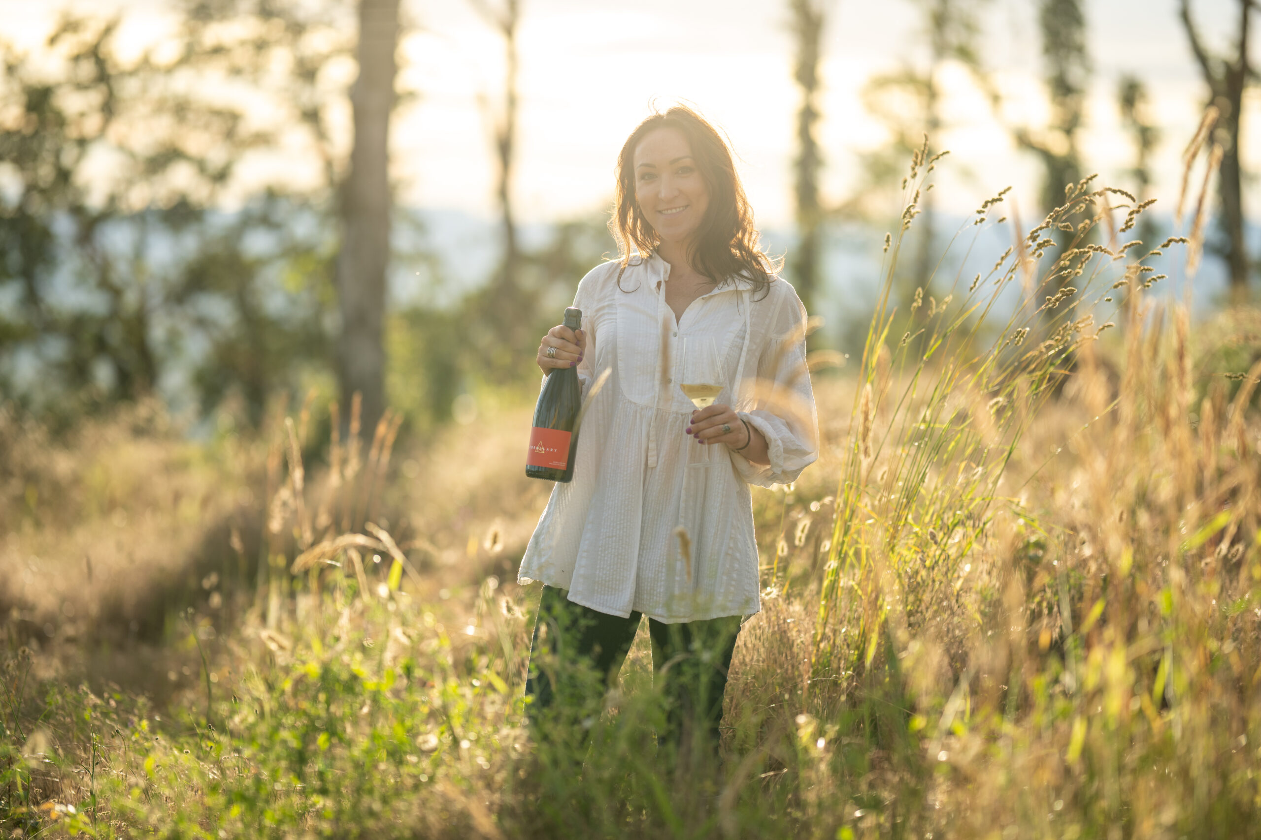 Woman in a white blouse with long brown hair standing in a sun filled field holding a bottle of wine.