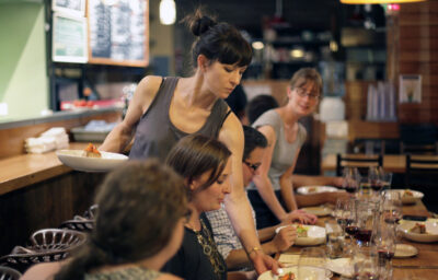 Woman serving food to people sitting at a long wooden table in a busy, well lit restaurant.