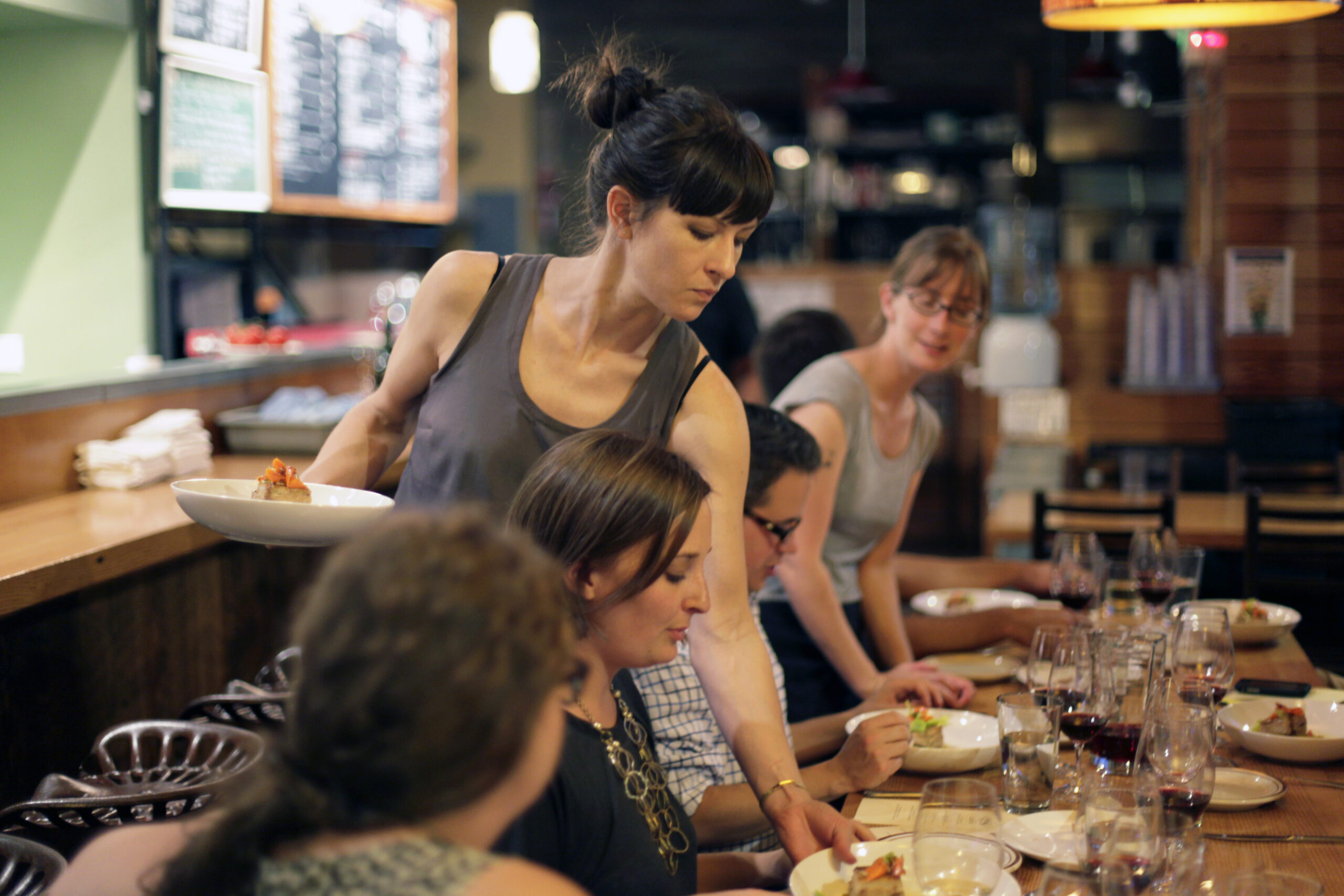 Woman serving food to people sitting at a long wooden table in a busy, well lit restaurant.