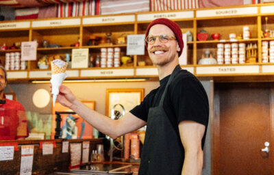 A person wearing a black shirt, black apron, glasses and red stocking cap serving an ice cream cone with vanilla ice cream standing at a counter.