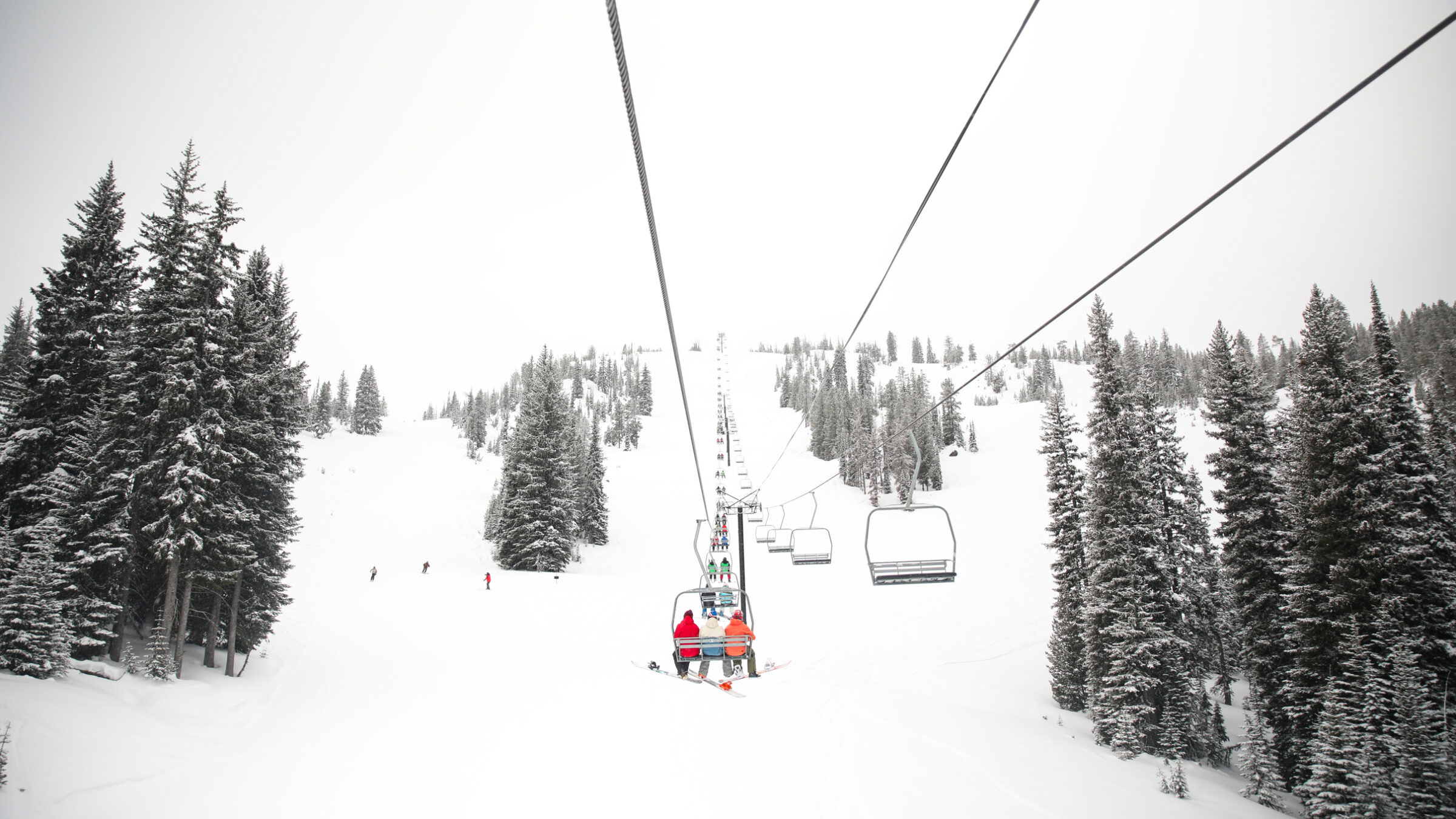 People riding a chair lift over a snowy mountain bordered by tall green trees and white/grey sky