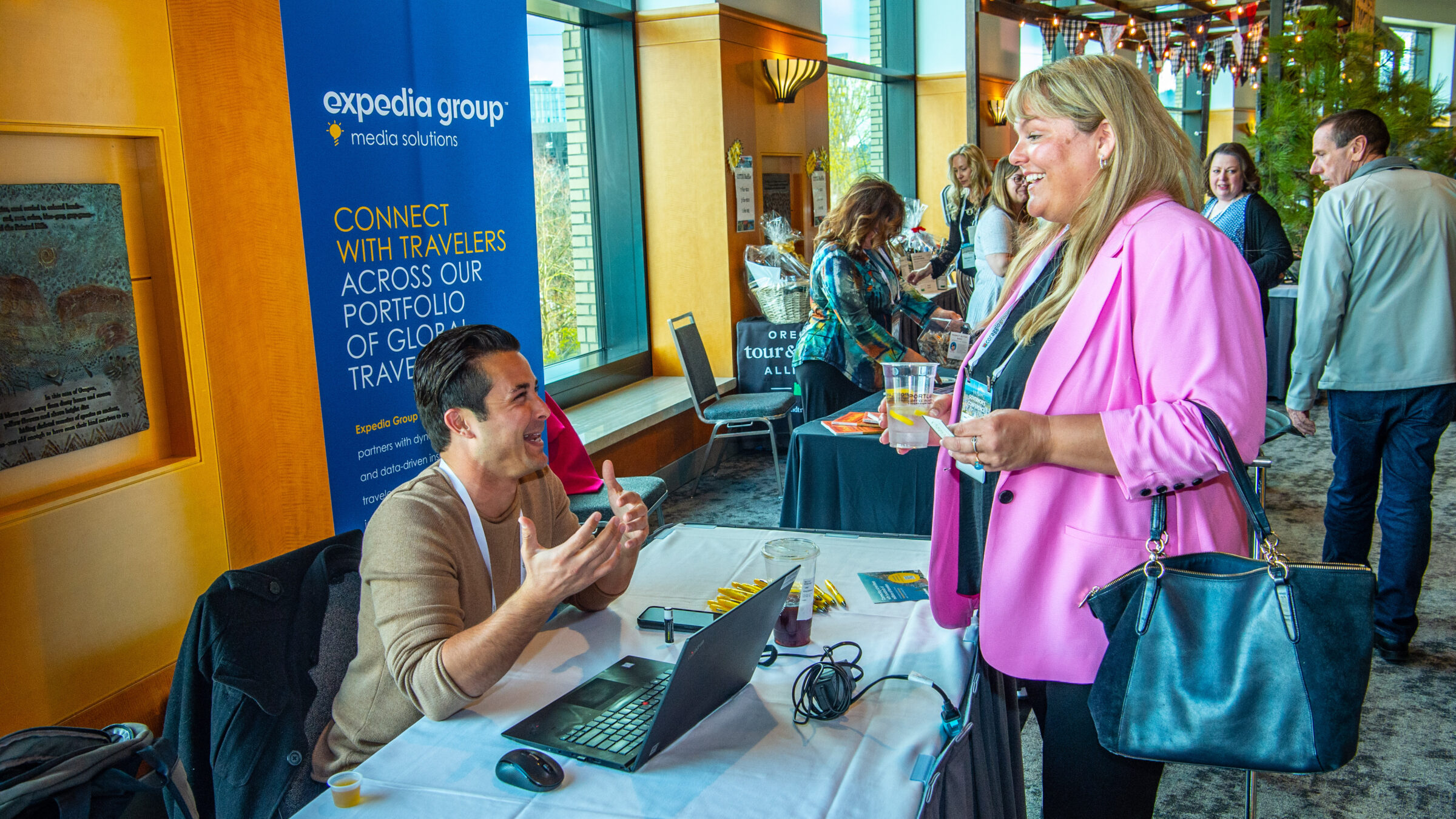 A person in a pink blazer standing at an exhibitor table talking to a person sitting at a table in the exhibitor hall of a conference