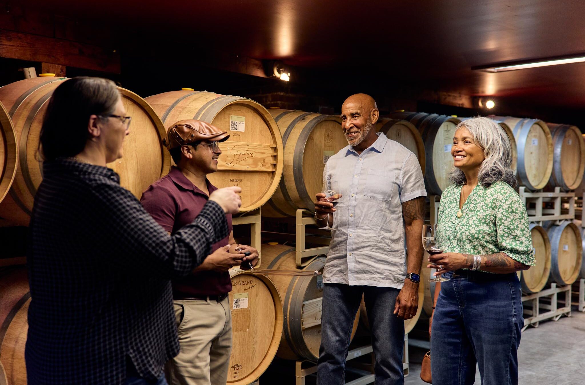 Four smiling people huddled inside a winery with wine glasses in hand and wood wine barrels behind them