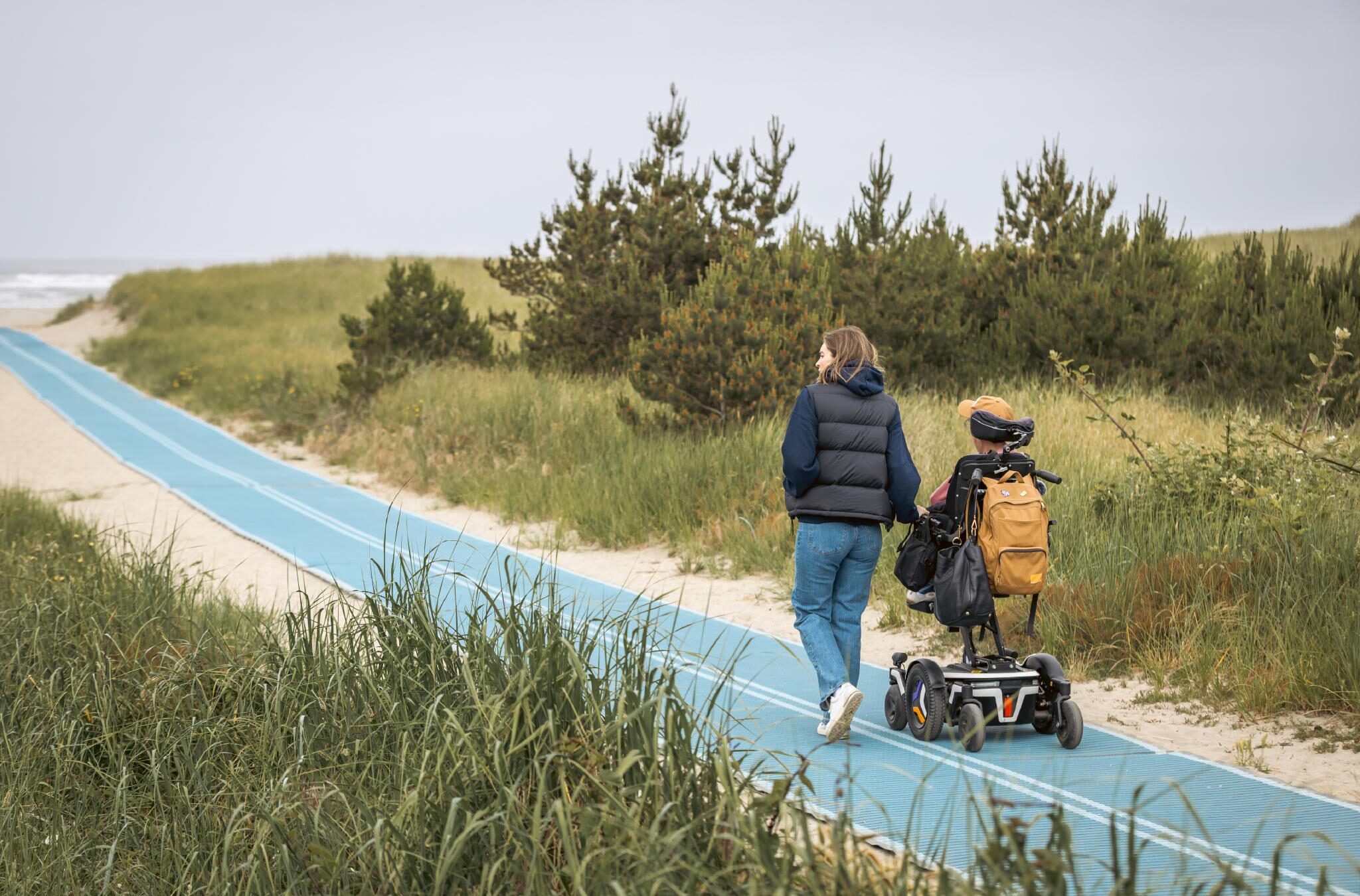A wheelchair user accompanied by a person walking, using a mobility mat to access the beach