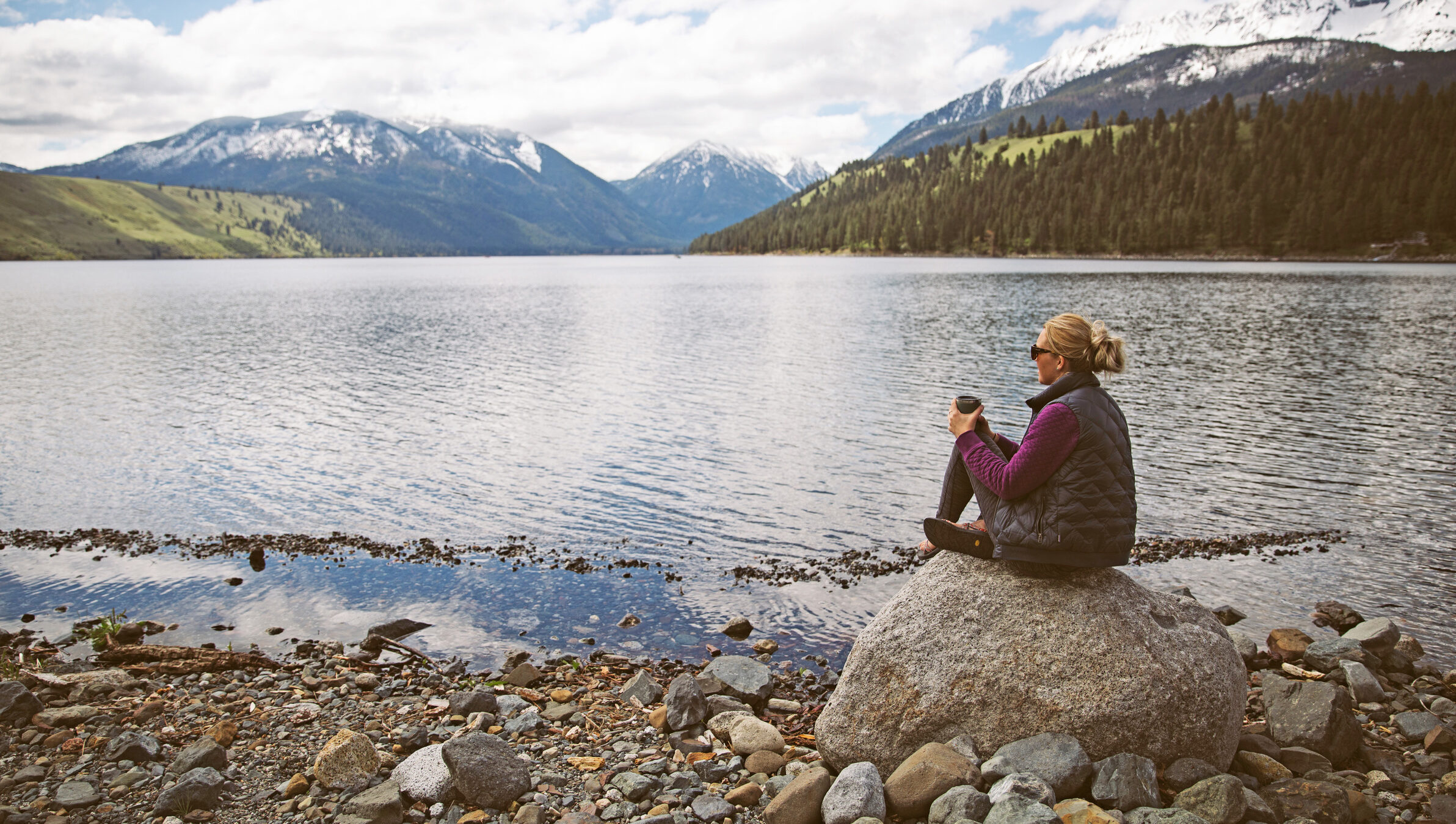 A woman sitting on a rock enjoying coffee and a view of a lake surrounded by snow capped mountains